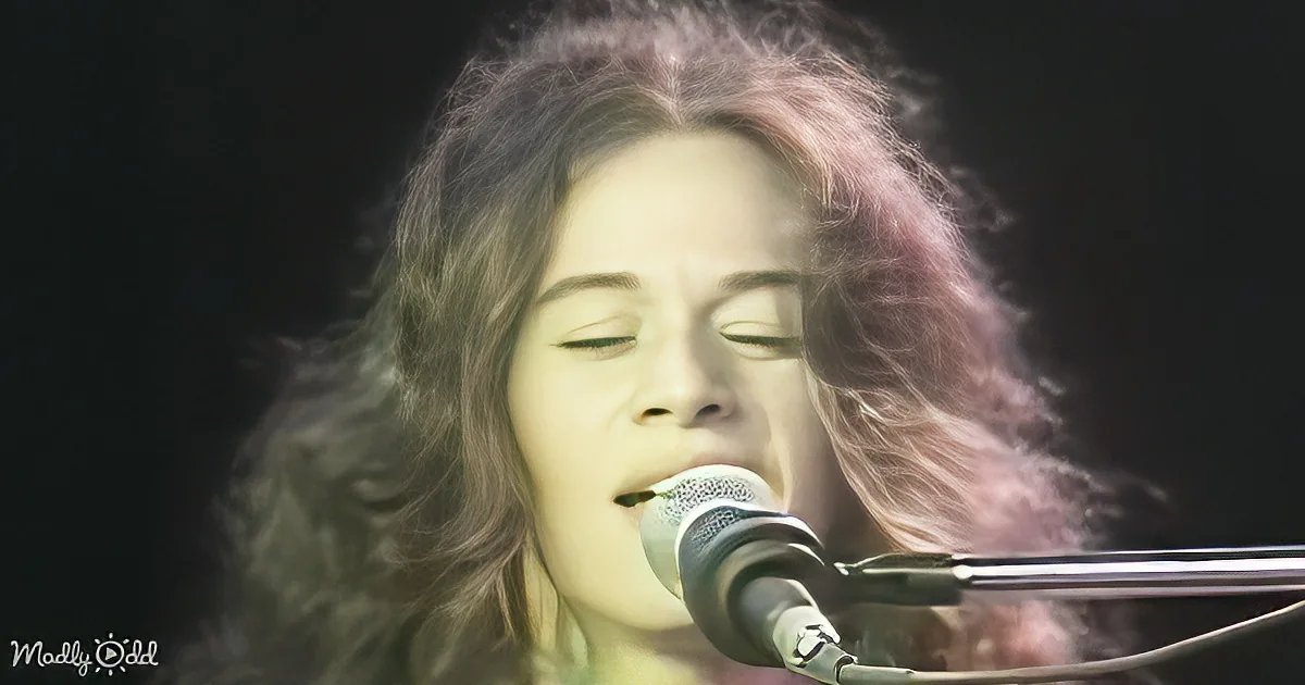 A person with long, wavy hair sings into a microphone, eyes closed, in a dimly lit setting.