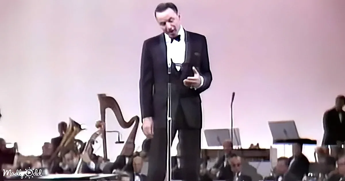 A man in a tuxedo sings on stage with an orchestra in the background.