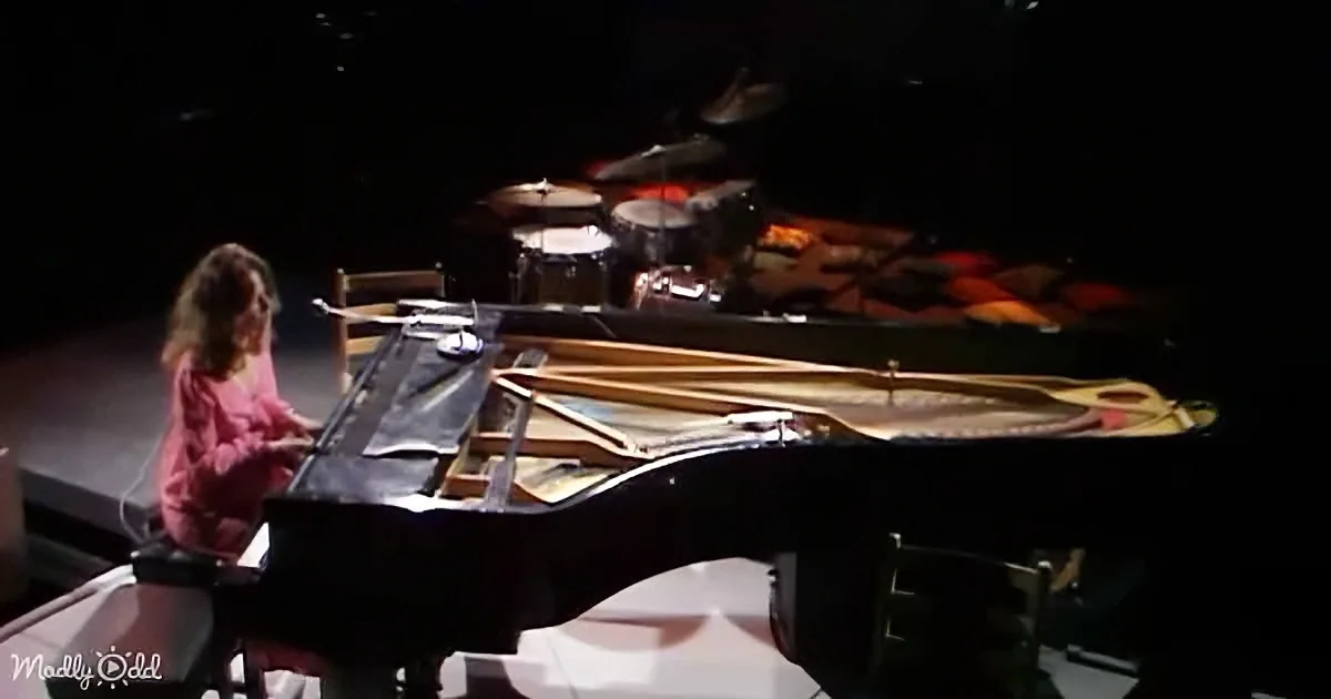 A young girl in pink plays a grand piano on stage, with drums and other instruments in the background.