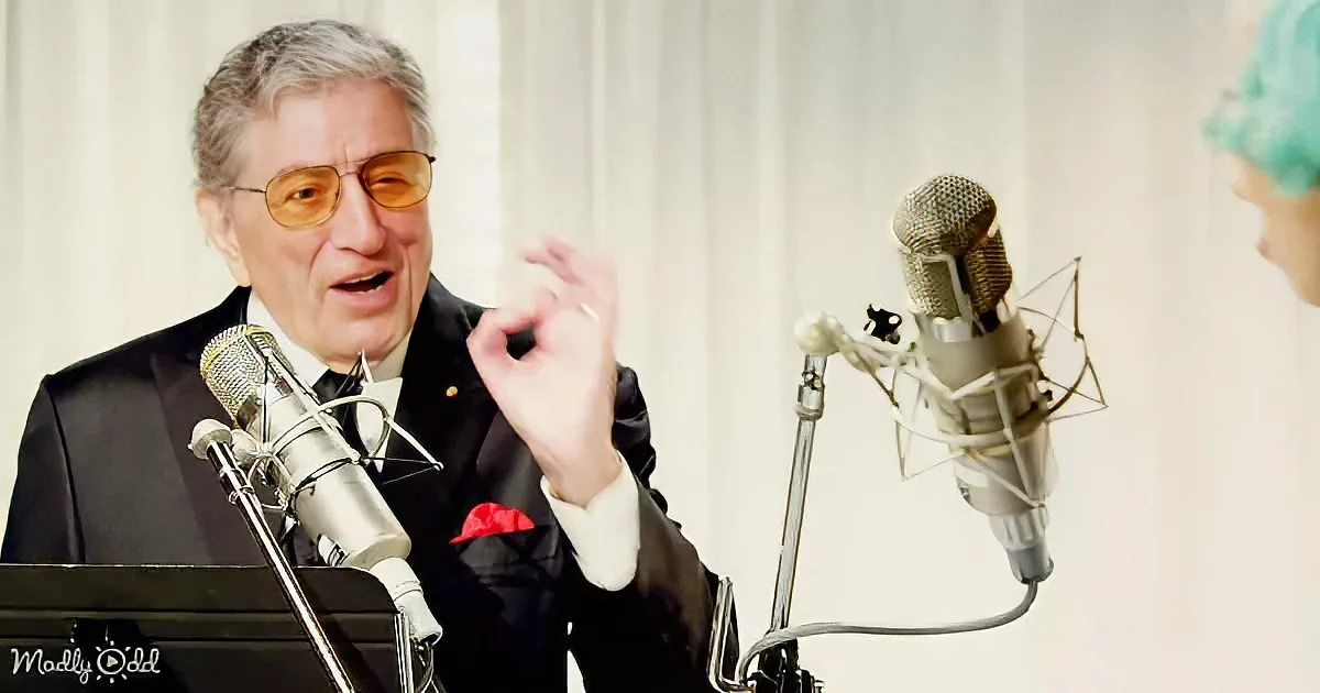 A man in a suit and glasses speaks into a microphone, gesturing with his hand.