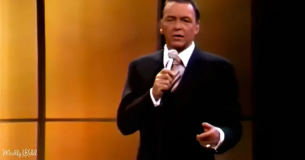 A man in a suit holds a microphone, gesturing with his other hand, against a warm-toned background.