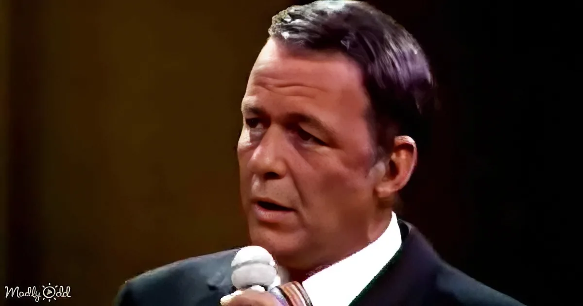 A man in a suit holds a microphone, speaking with a serious expression against a dark background.
