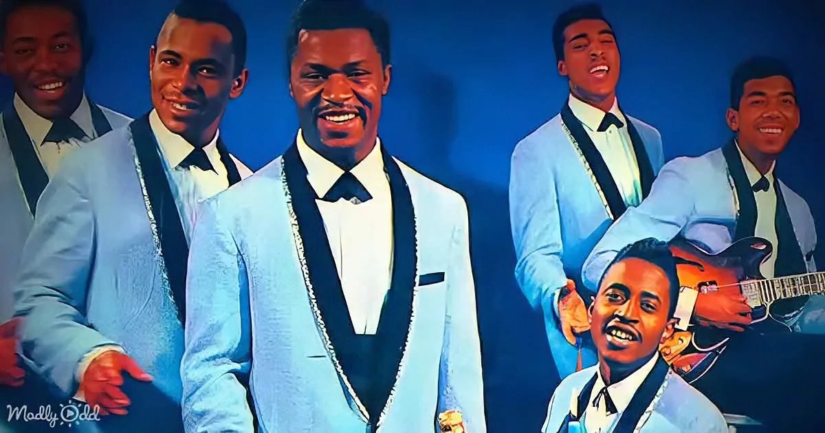 A man in a light blue suit and black bow tie smiles, surrounded by other men in similar attire, one playing guitar.