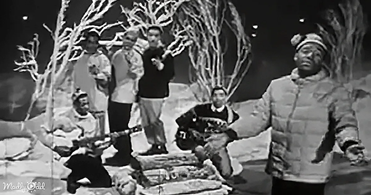 A man in a winter coat stands with others in a snowy scene, holding a rifle.