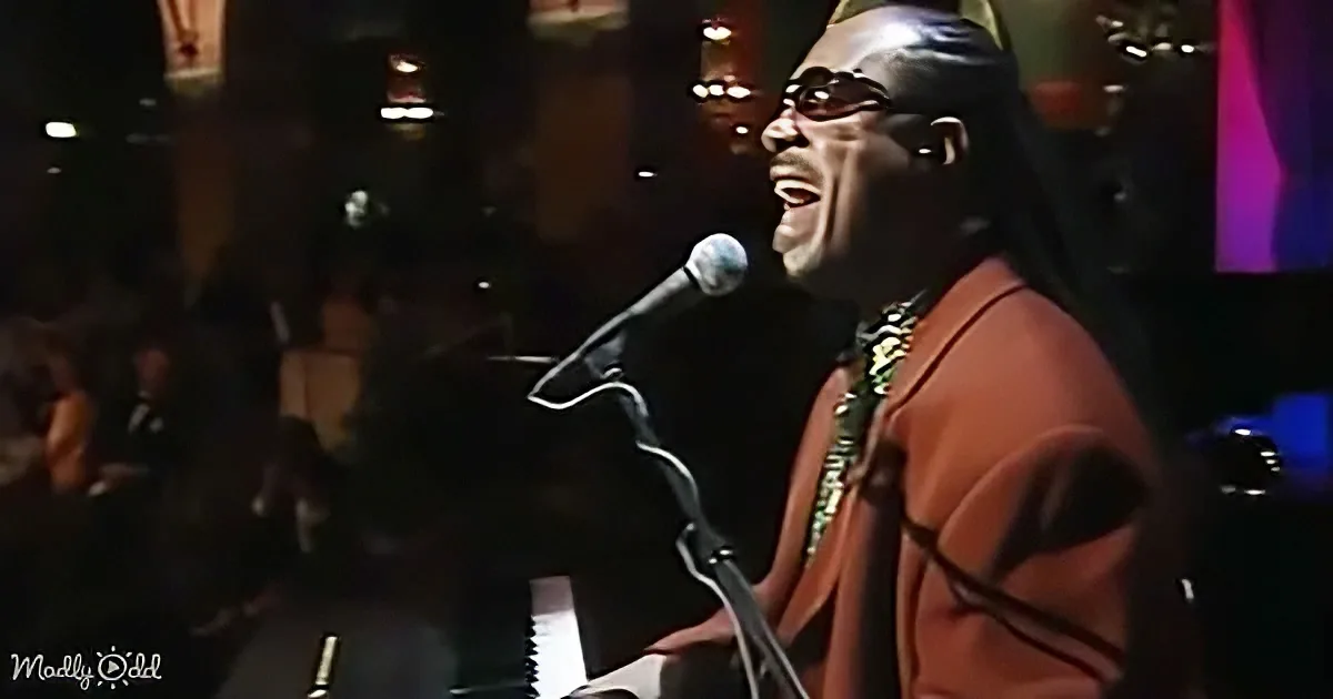A man in a red suit sings into a microphone on stage, with a dark background and audience.