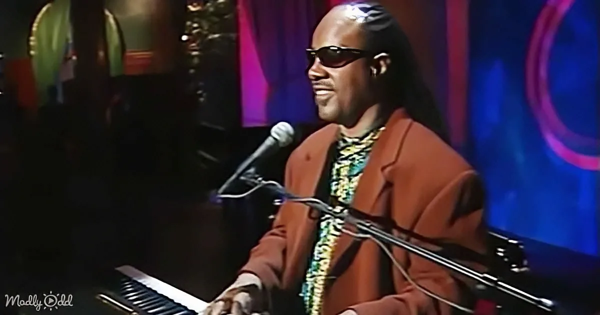 A man in sunglasses and a brown jacket plays piano on a stage with a microphone and colorful background.