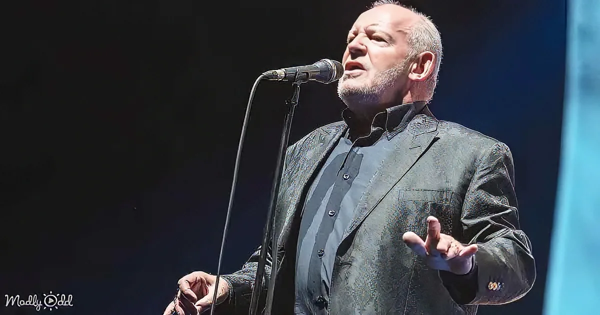 A man in a suit sings into a microphone, gesturing with his hands.