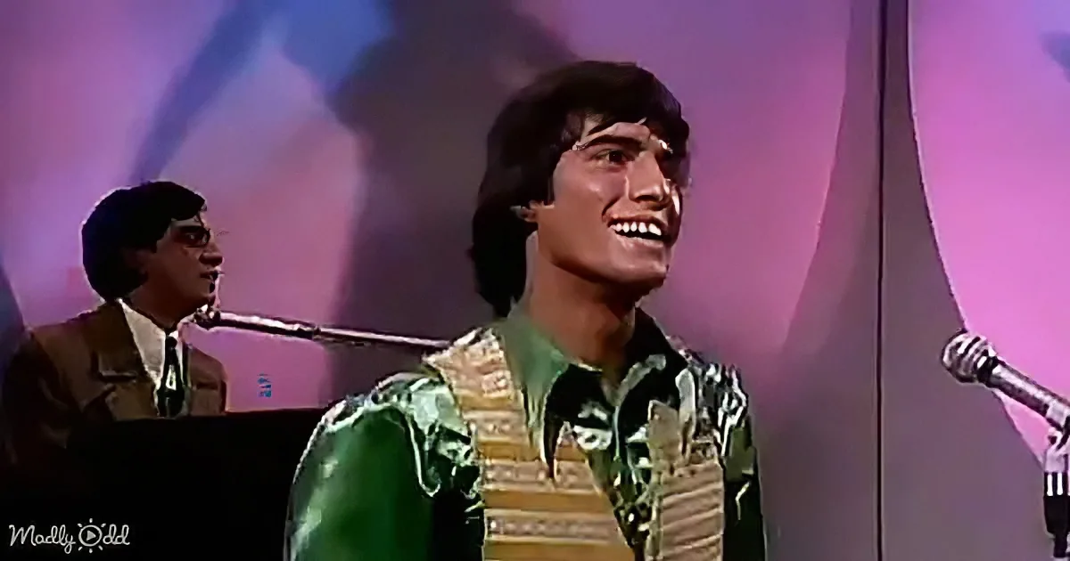 A man in a green and yellow patterned shirt smiles on stage with a keyboard player in the background.