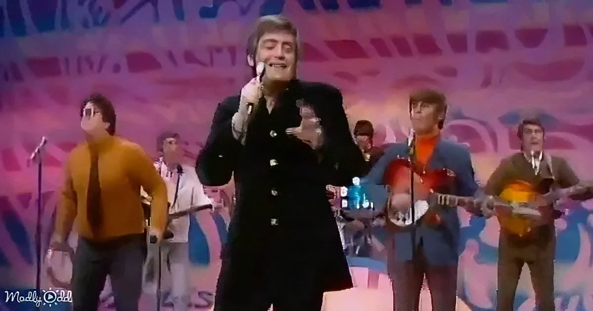 A man in a black suit sings into a microphone on stage, surrounded by musicians.
