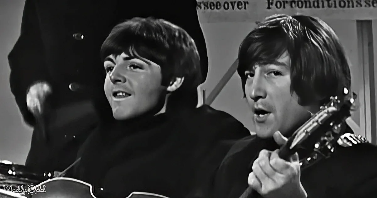 Two individuals, one playing guitar, both in black, singing and smiling, in a black-and-white setting.