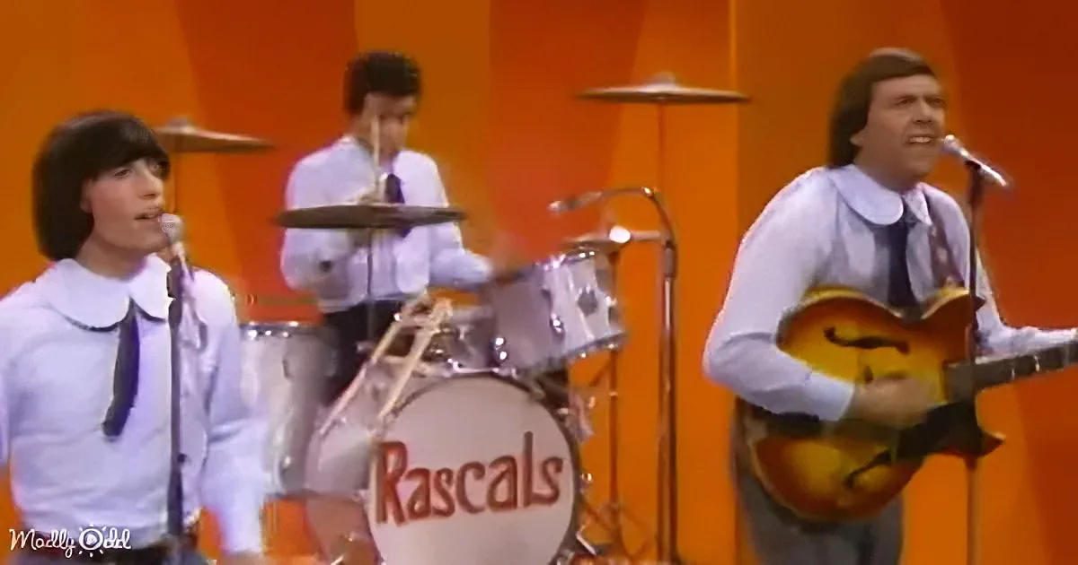 A band in white shirts and ties performs on stage, with a drummer and guitarist visible.