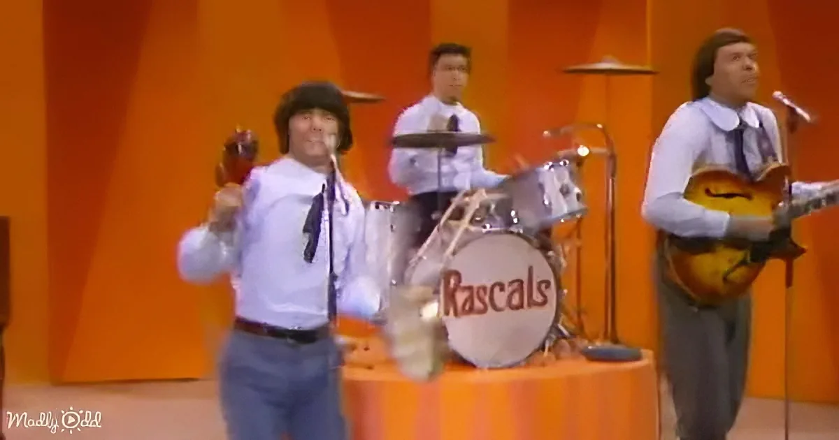 A man in a white shirt and tie sings into a microphone on stage with a band.