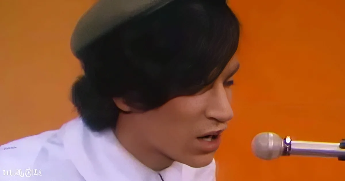 A person wearing a beret and white shirt sings into a microphone against an orange background.