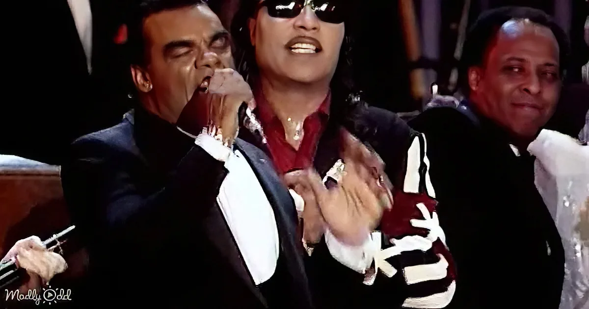 A man in a suit is passionately singing into a microphone, surrounded by other people.