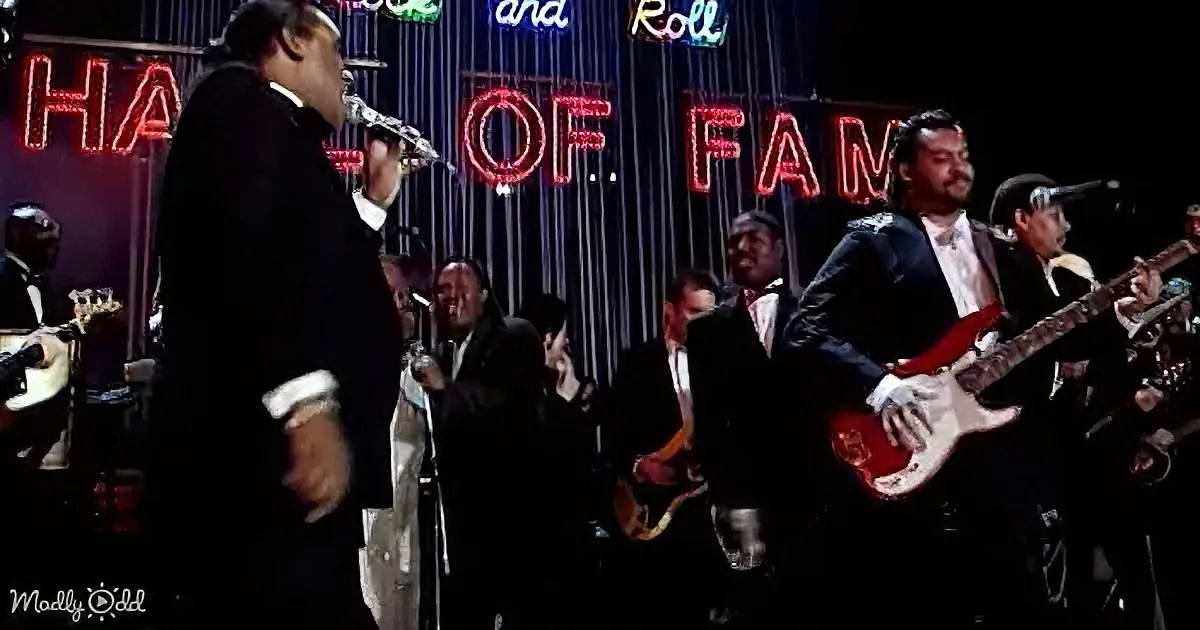 A man in a suit sings into a microphone on stage with a band, "Rock and Roll Hall of Fame" sign in the background.