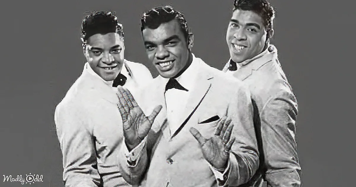 Three smiling men in suits, one in a white coat, posing together with hands raised.