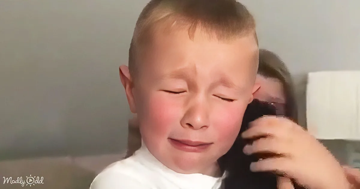 A young boy with short hair, wearing a white shirt, is crying while being held by someone.