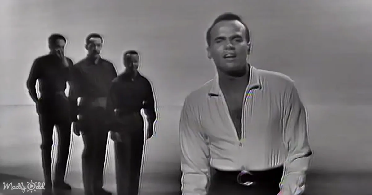 A man in a white shirt sings in the foreground, with three other men in dark suits standing behind him.