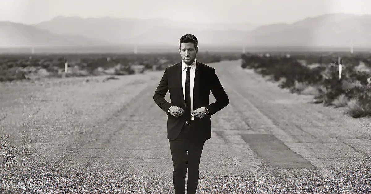 A man in a suit stands on a desert road, buttoning his jacket, with mountains in the background.