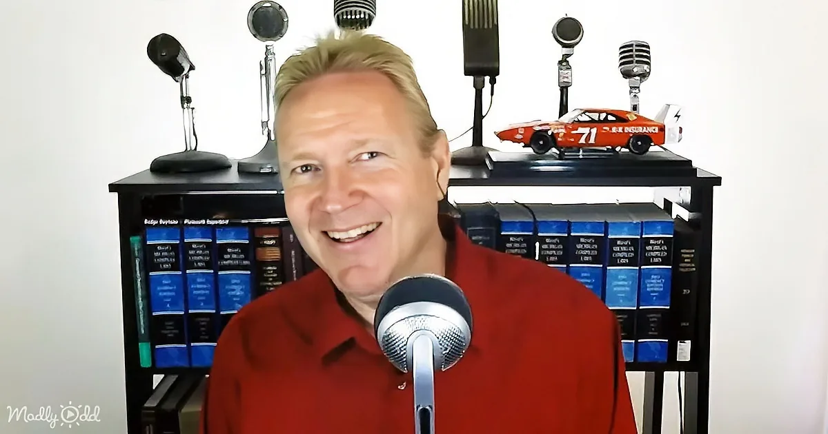 A man in a red shirt smiles in front of a microphone, with a bookshelf and model car in the background.