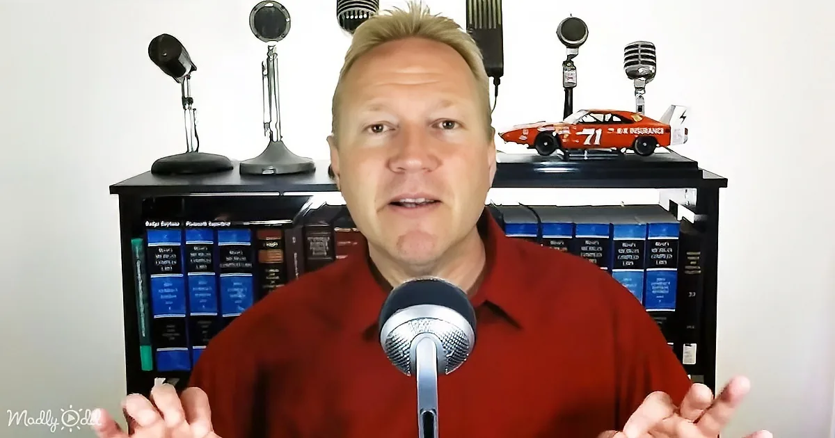 A man in a red shirt speaks into a microphone, gesturing with his hands, with a bookshelf and model car in the background.