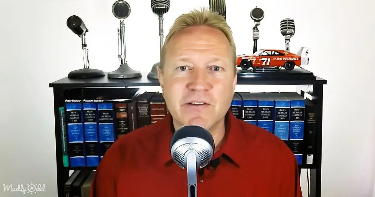 A man in a red shirt speaks into a microphone in front of a bookshelf with microphones and a toy race car.