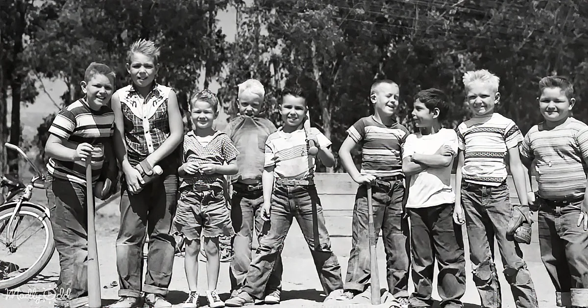 A group of boys in casual attire stand outdoors, some holding tools, with trees and a bicycle in the background.