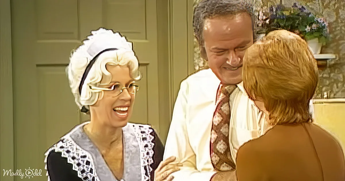 A woman in a maid's outfit and a man in a suit are smiling at each other, with another woman in a beige top nearby.