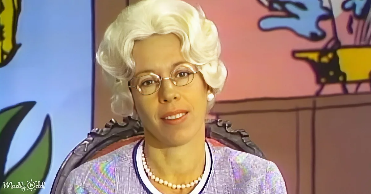 A woman with white hair, glasses, pearls, and a purple outfit sits in a chair against a colorful background.