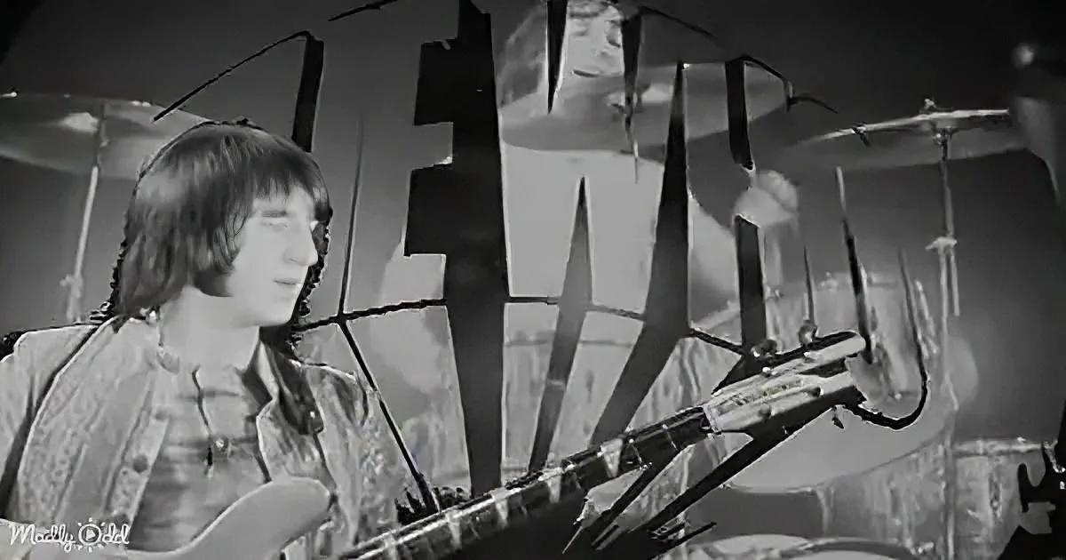 A musician plays guitar in front of a large "FAT" sign, with a drum set in the background.