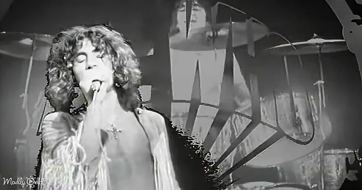 A singer with curly hair, wearing a white outfit, passionately sings into a microphone on stage.
