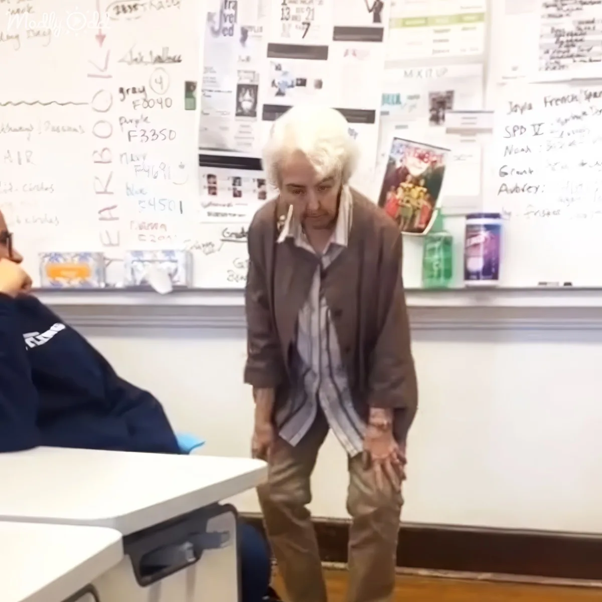 An older man with white hair stands in a classroom, leaning forward with hands clasped, near a whiteboard filled with...