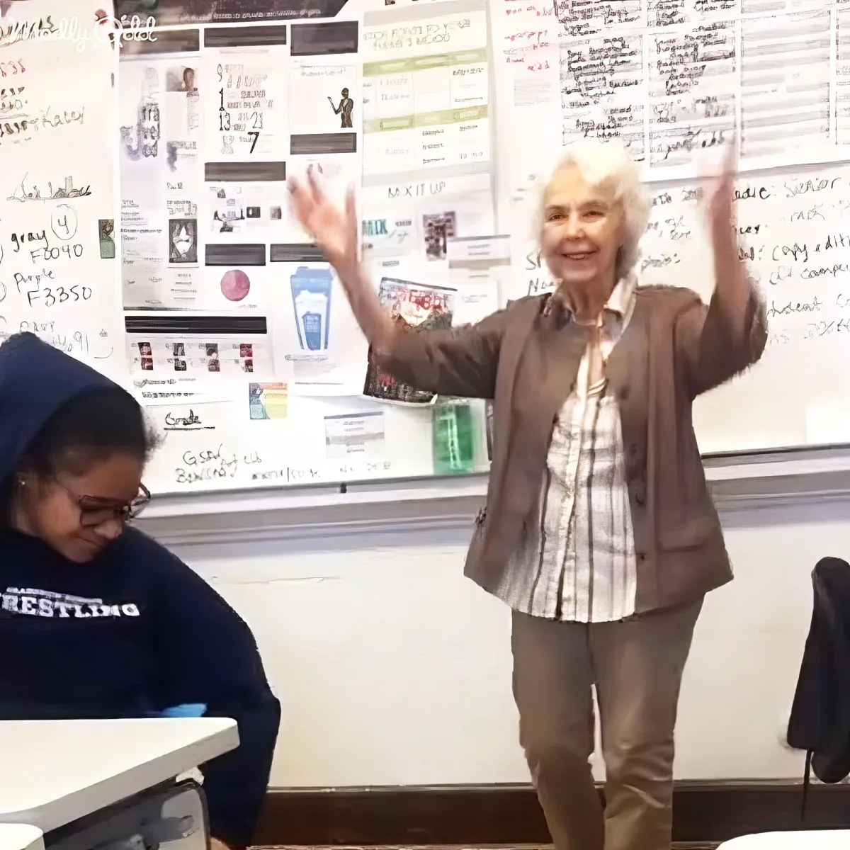 An older woman stands in a classroom with her arms raised, smiling, while a student sits at a desk nearby.
