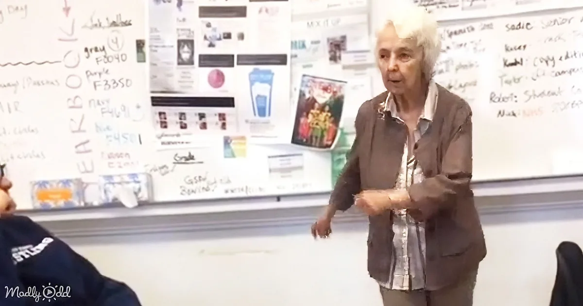 An older woman stands in front of a whiteboard, gesturing while speaking in a classroom setting.