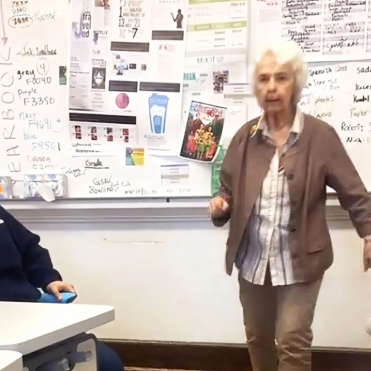 An older woman stands in front of a classroom, gesturing, with a whiteboard behind her covered in notes and colorful papers.