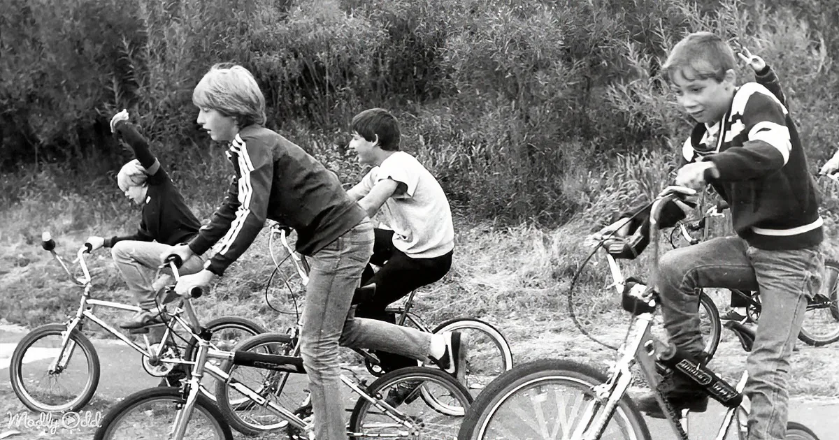 Four boys riding bicycles in a line, one making a peace sign, black and white photo.
