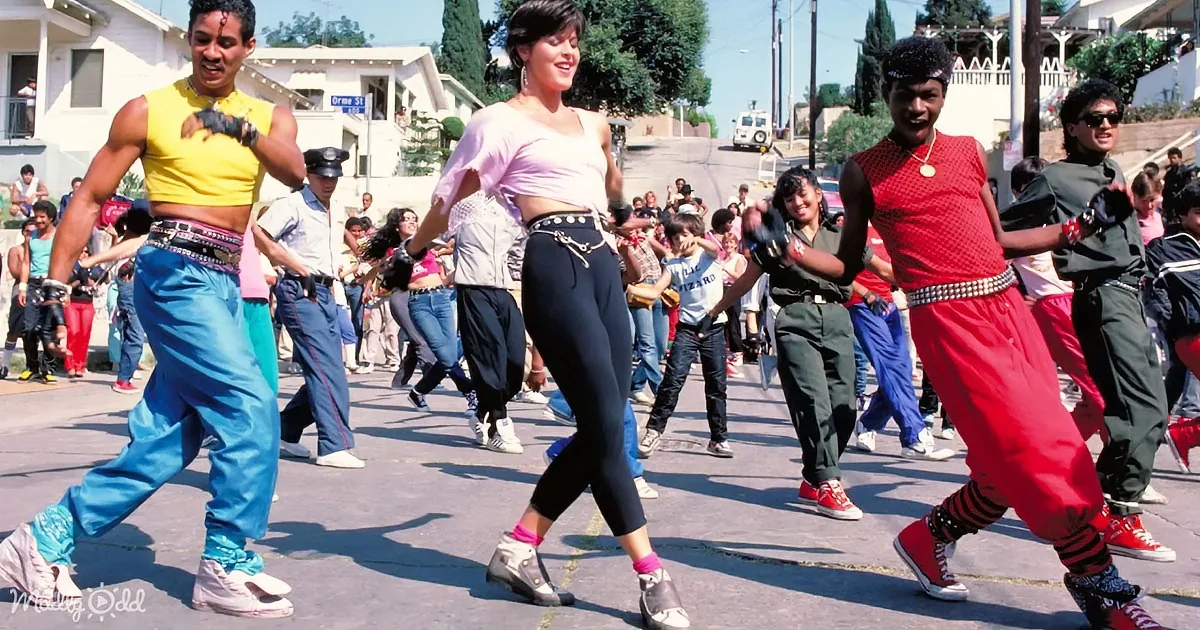 A group of people in vibrant 80s attire dances energetically on a sunny street.
