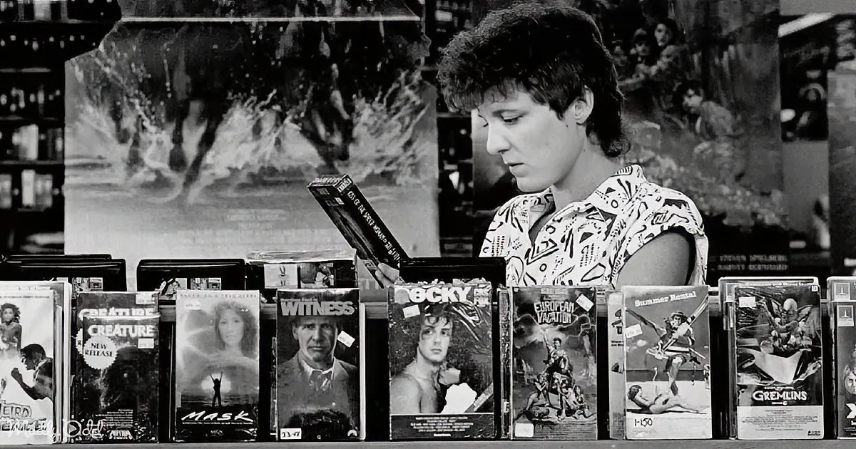 A person examines a VHS tape in a video rental store surrounded by movie cases.