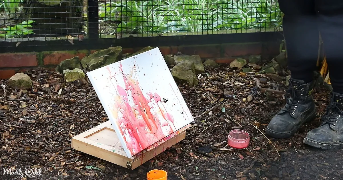 A canvas with red and pink paint splatters sits on an easel outdoors, surrounded by mulch and a person in boots.