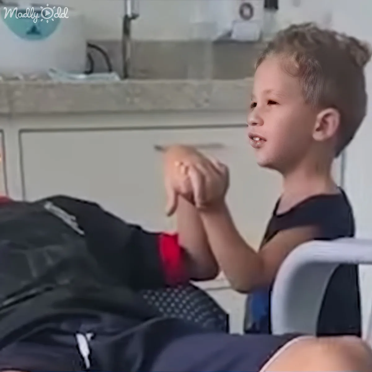 A young boy with blonde hair sits in a chair, holding a black backpack with a red strap.
