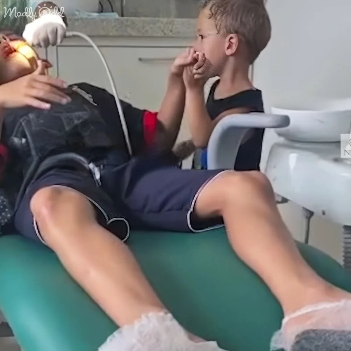 A young boy sits in a dental chair, covering his face, while a dentist works on his teeth.