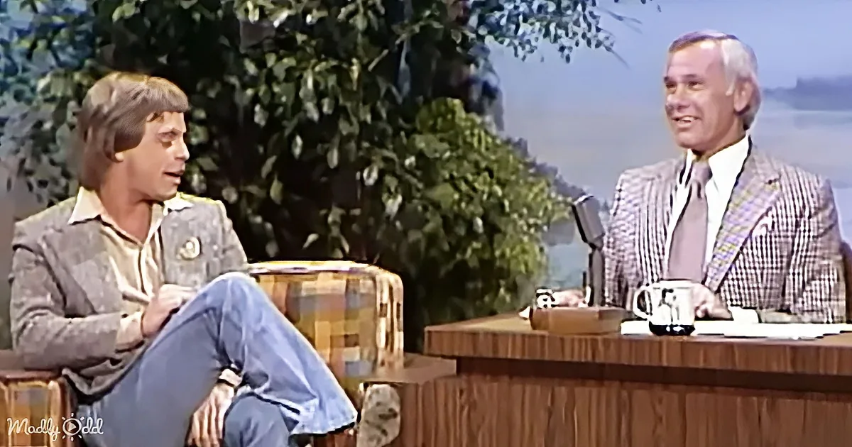 A man in a plaid suit sits at a desk, smiling, while another man in casual attire sits on a plaid chair, appearing to talk.
