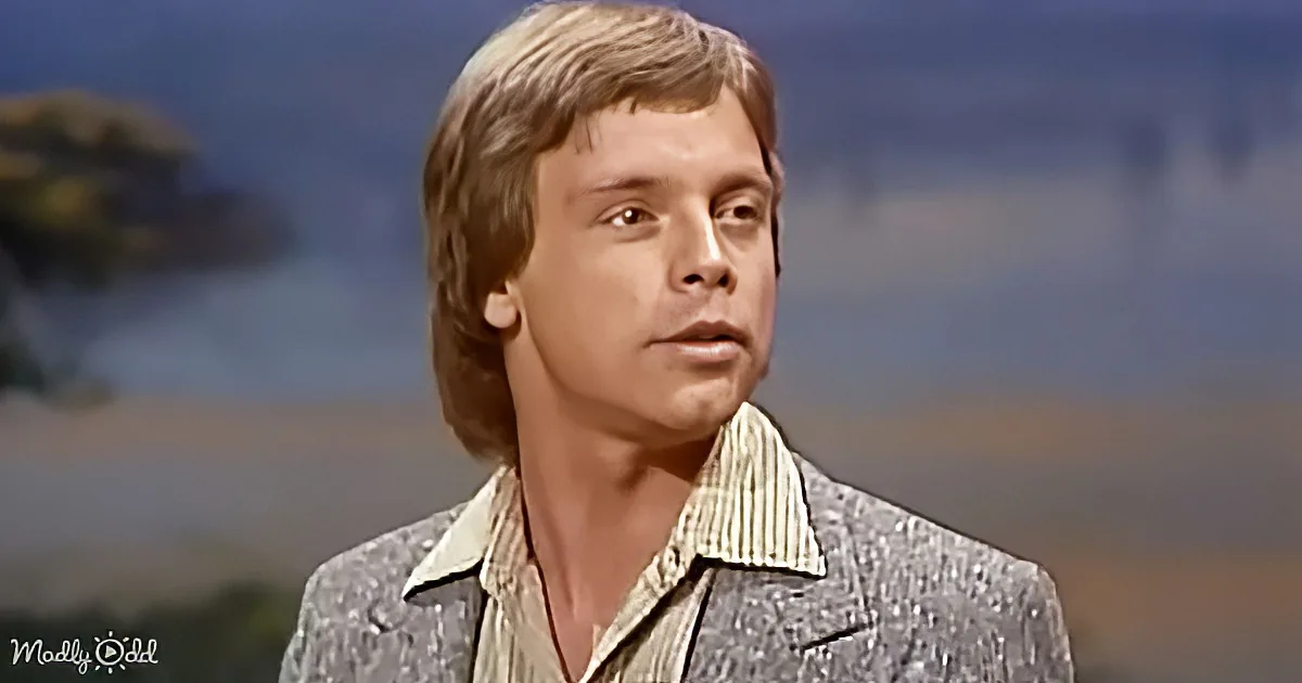 A man with shoulder-length hair, wearing a patterned blazer and striped shirt, stands against a blurred background.