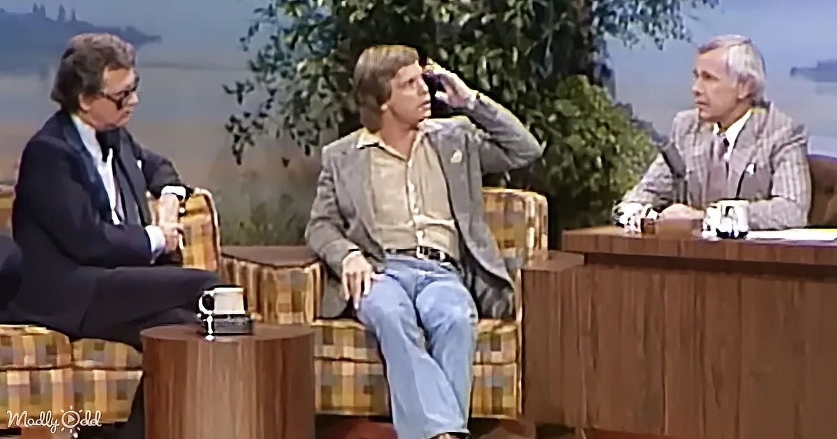 A man in a suit sits on a plaid chair, gesturing with his hand, while another man in casual attire sits beside him, and a...