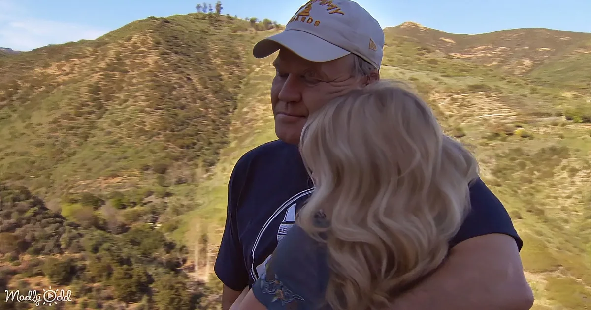 A man in a cap and blue shirt embraces a woman with blonde hair, set against a scenic mountain backdrop.