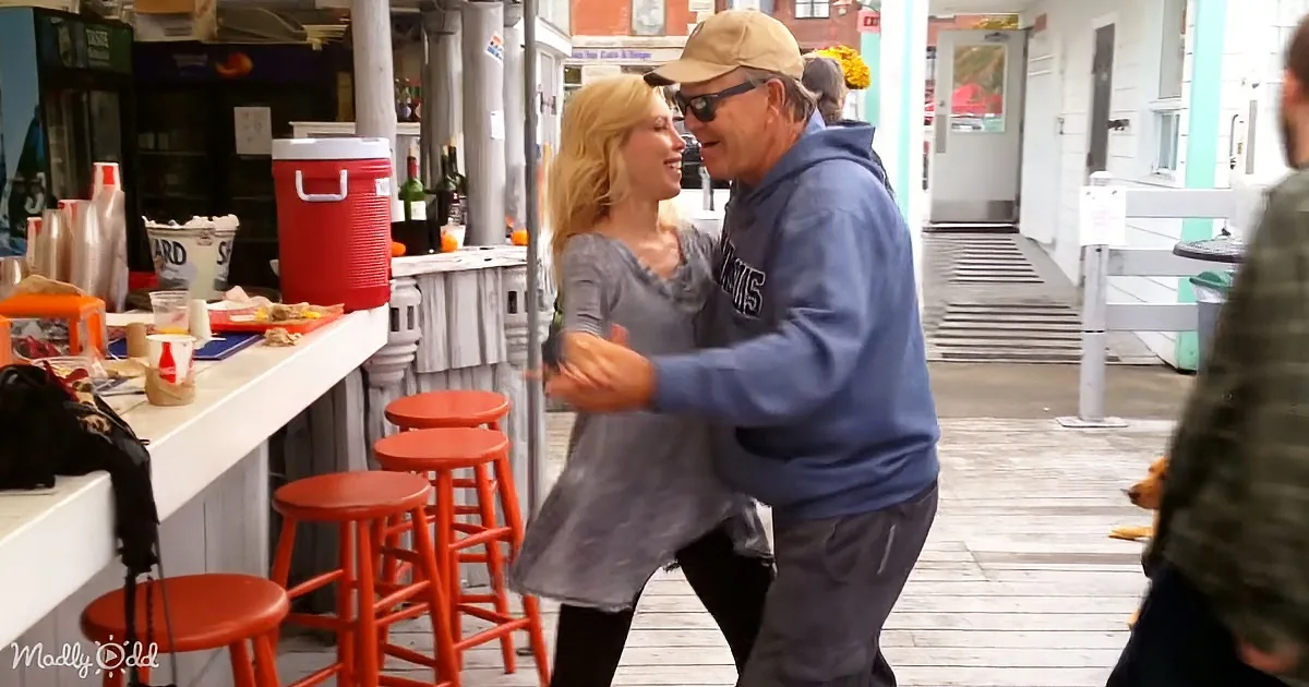 A man and woman are dancing closely, smiling, in front of a bar with red stools and a red cooler.