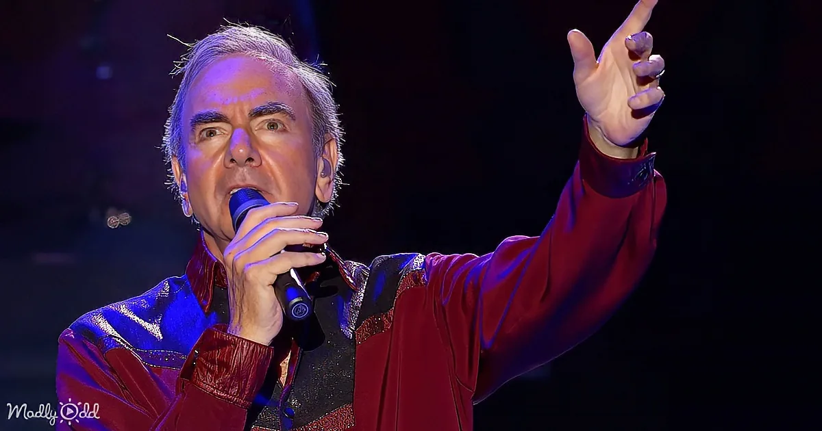 A man in a red and black outfit sings into a microphone, pointing upwards with his right hand.
