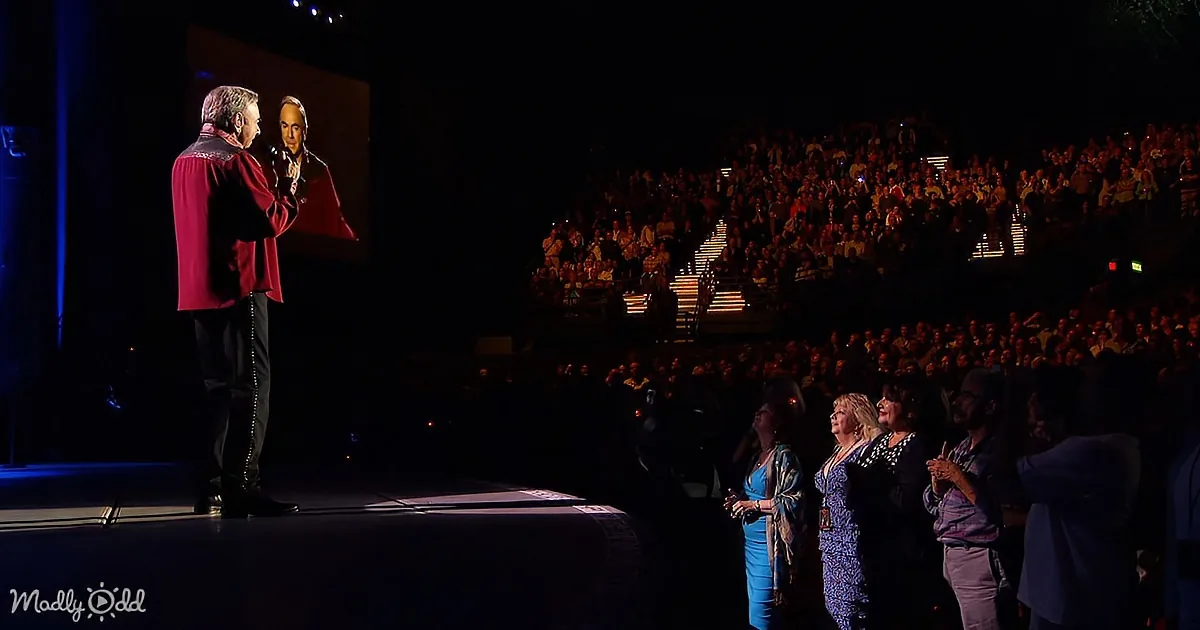 A man in a red cape stands on stage, addressing a large audience in a dimly lit auditorium.