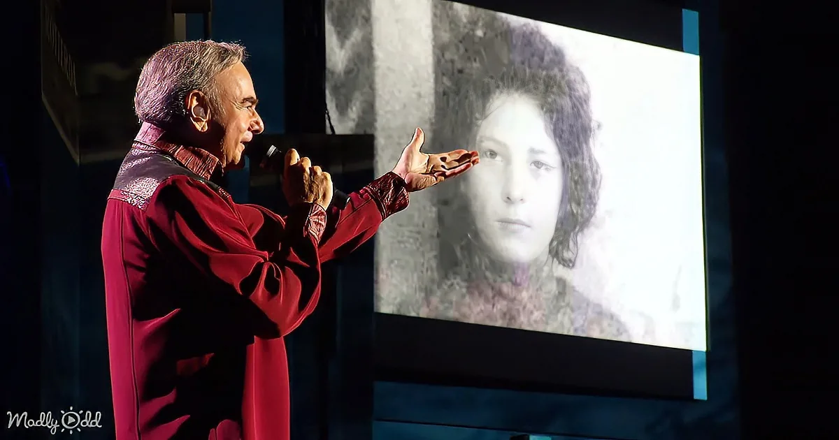 A man in a red robe gestures while speaking, with a projected image of a person behind him.