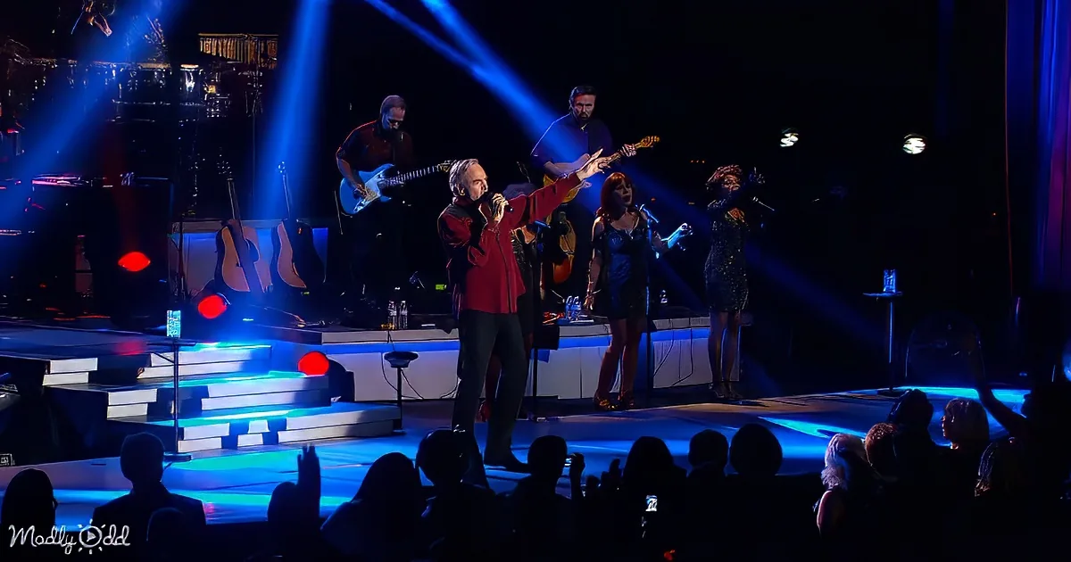 A man in a red shirt sings on stage with a band, illuminated by blue lights, while an audience watches.